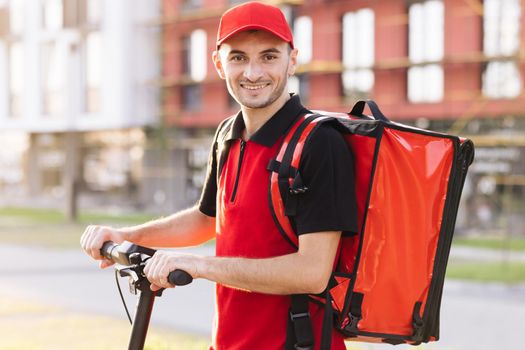 Male courier with red backpack and electric scooter. Delivering green transportation. Portrait shot of man delivery worker in red uniform standing at street and turning face to camera.