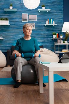Senior woman exercising body muscles doing arms exercise sitting on swiss ball in living room. Pensioner watching online aerobics training on tablet exercising muscular resistance holding dumbbell