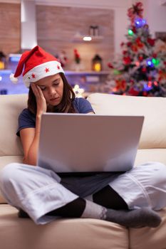 Tired exhausted adult falling asleep while reading business email on laptop computer sitting on couch in xmas decorated kitchen. Woman enjoying winter holiday celebrating christmas season