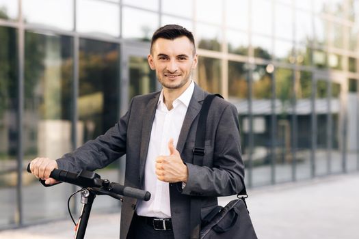 Handsome businessman with his electric scooter in front the business building. Young businessman in elegant suit standing outdoors, cheerfully smiling and showing thumb up.