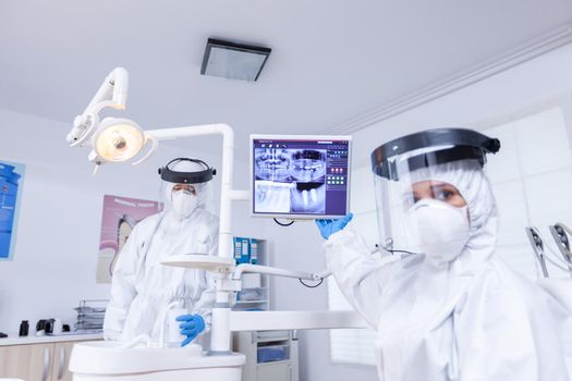 Patient pov of dentist with face shield explaining dental x-ray pointing at monitor. Stomatology specialist wearing protective suit against infection with coronavirus pointing at radiography.
