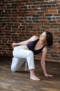 Beautiful young woman with white sporty pants and black top practicing hatha yoga Parivrtta Anjaneyasana pose crescent lunge on knee with twist, against the background of a brick wall in the loft.