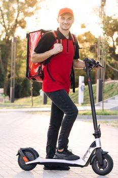 Portrait of deliverman standing with electric scooter with red thermal backpack looking at camera. E-Scooter rider eco transport. Modern urban alternative transport. Excellent delivery.
