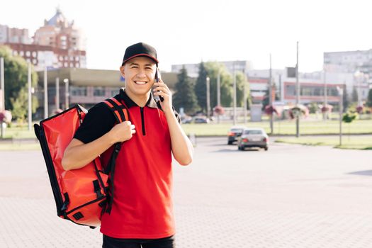 Male courier with isothermal food case box arrives to the entrance to the house and calls for client. Food delivery guy with red backpack deliver orders.