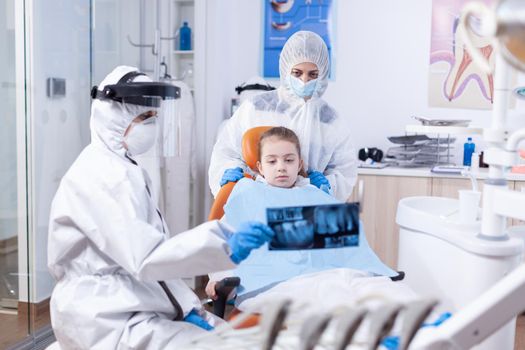 Orthodontist in ppe suit using dental radiography to explain dentistiry procedure for mouth hygine. Stomatolog in protectie suit for coroanvirus as safety precaution holding child teeth x-ray during consultation.