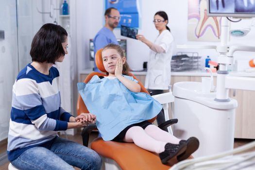 Little girl in dentist office showing parent where tooth hurts touching face with painfull expression. Child with her mother during teeth check up with stomatolog sitting on chair.