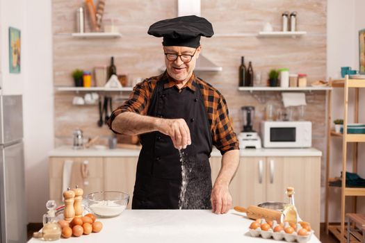 Hand spreading wheat flour on wooden kitchen table for homemade pizza. Retired senior chef with bonete and apron, in kitchen uniform sprinkling sieving sifting ingredients by hand.