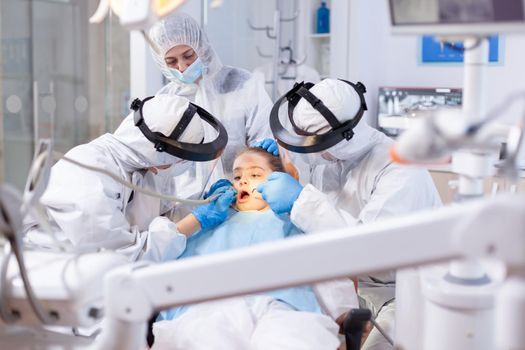 Orthodontist and assistant treating little girl caries dressed in ppe suit as safety precaution against coronavirus outbreak. Stomatology team wearing ppe suit during covid19 doing procedure on child teeth.