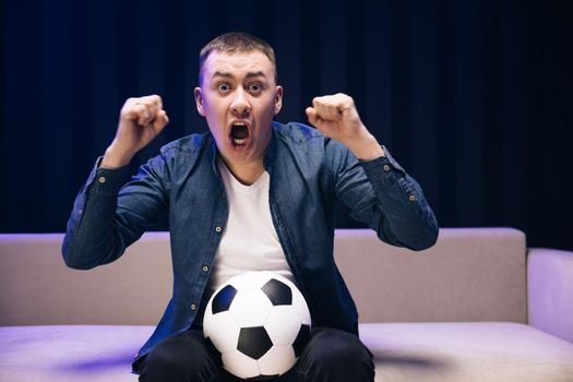 Close up of man with ball in hands watch sport channel. Handsome Caucasian man worried and cheering up for football game on TV sitting on sofa late at night.