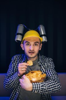 Hipster man with beer helmet on the head eating chips while watching TV At home.