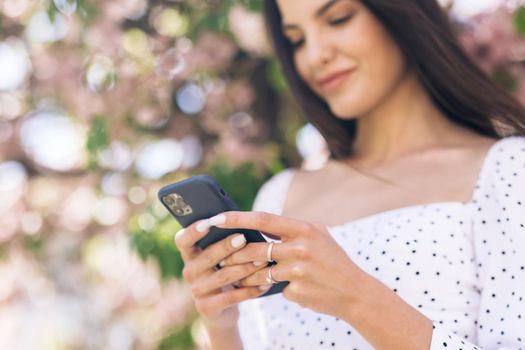 Portrait of happy hipster woman typing by mobile phone outdoors. Cheerful girl walking with smartphone in urban background. Smiling lady holding cellphone in hands outside.