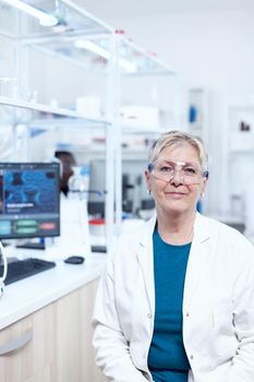 Smiling senior chemist wearing protective glasses with test tubes behind. Elderly scientist wearing lab coat working to develop a new medical vacine with african assistant in the background.