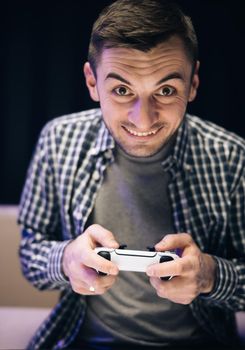 Man in casual wear playing video game in home living room. Caucasian guy holding joystick control having fun laughing on couch indoors.