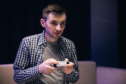 Caucasian guy playing video game and using joystick . Handsome young man enjoying free time alone while sitting on couch in living room.