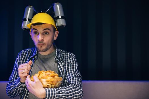 Man with beer helmet on the head eats chips while sitting at home on couch, in evening he watches movies at home on TV. Caucasian man rests at home and watches TV shows or sports news on TV screen.