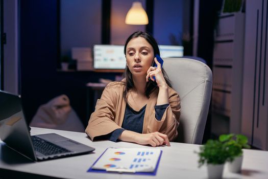 Workaholic manager talking with customer over the phone in the evening. Woman entrepreneur working late at night in corporate business doing overtime in the course of phone call.