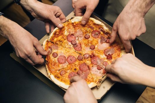 Group of hungry friends sitting at desk and sharing delicious lunch on table background. Male hands taking slices of pizza with cheese, tomatoes and ham from food delivery.