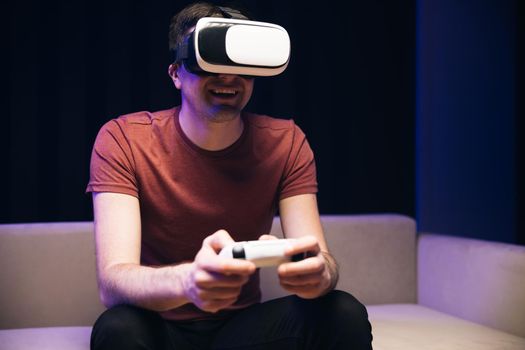 Attractive caucasian man hold joystick and wear virtual reality glasses headset helmet on sofa at home. Gamer playing game using VR glasses at home.