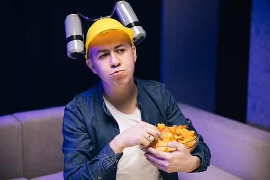 The guy with beer helmet on the head sits on the couch, eats chips and watches the TV. Handsome cute home interior technology portrait