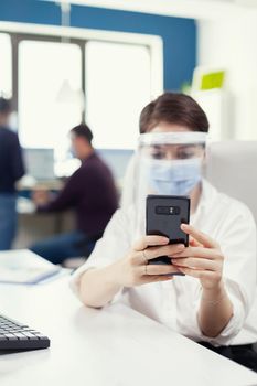 Businesswoman using mobile phone texting on smartphone wearing face mask as safety precaution against covid19. Multiethnic coworkers working respecting social distance in financial company.