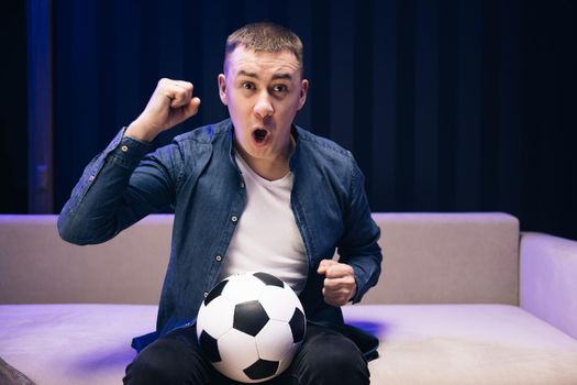 People emotions sport leisure lifestyle concept. Young fun guy 25s football fan cheer up support favorite team hold soccer ball in denim jacket white t-shirt in dark living room.