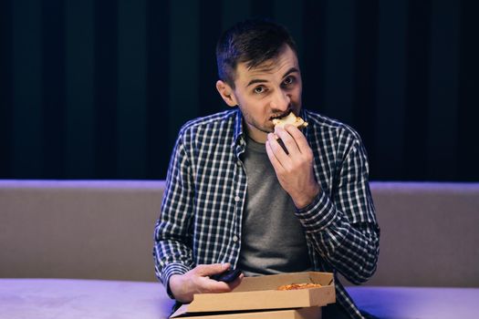 Handsome young man eating pizza while watching tv while relaxing at sofa in the living room.