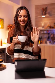 Young adult holding cup of tea while waving on online chatting video call conference with friends. Caucasian woman using wireless internet technology on tablet in christmas decorated home