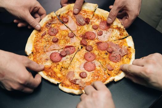 Top view male hands taking slices of pizza with cheese, tomatoes and ham from food delivery. Group of hungry friends sitting at desk and sharing delicious lunch on table background.