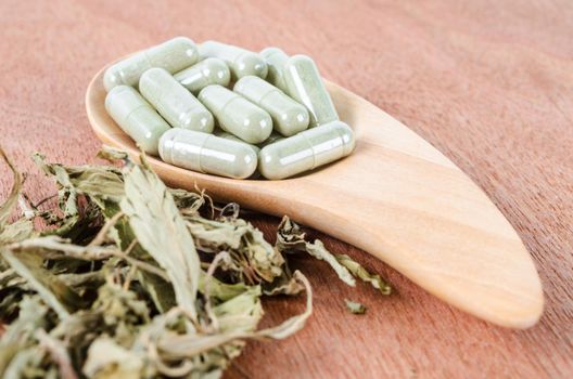 Pile of herbal medicine capsules in wooden spoon with dried leaves on wooden background.