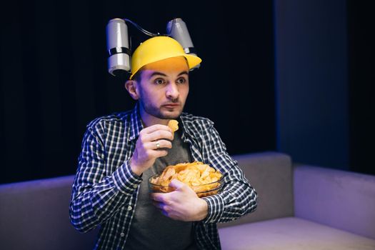 Guy with beer helmet on the head sits on the couch, eats chips and watches the TV. Handsome cute home interior technology portrait