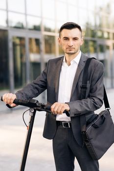 Stylysh man on vehicle outdoors. Modern urban alternative transport. Portrait of confident businessman standing with electric scooter and looking at camera. E-Scooter rider rent personal eco transport