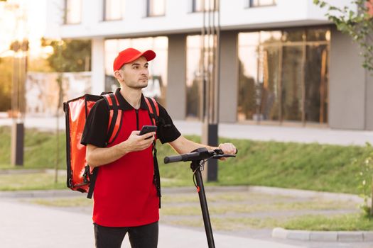 Male courier food delivery with red thermal backpack walks street with electric scooter uses smartphone navigate. Deliveryman worker employee deliver online order client customer at sunset