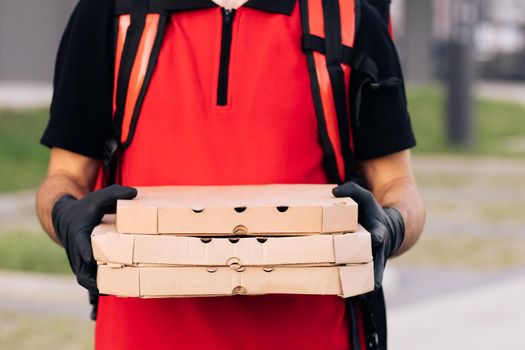 Portrait of handsome caucasian delivery man in gloves and medical mask standing at street, holding and handing carton boxes. Male courier giving pizza.