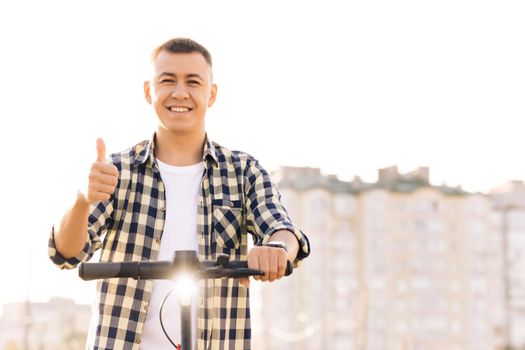 Caucasian hipster man leaning on electric scooter looking at camera and showing thumb up. Hipster man on vehicle outdoors. Eco-friendly modern urban transport. Ecology and urban lifestyle.