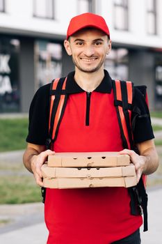 Portrait of young courier delivery man with red backpack holding pizza in carton boxes, waiting for customer in residential area. Delivery services. Excellent delivery.