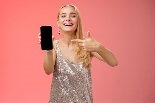 Boastful proud charming elegant blond woman in stylish evening dress show smartphone display proudly pointing mobile phone screen smiling showing photo boyfriend, standing red background.