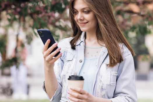 Attractive young woman with smartphone and cup of coffee in hands on background of sakura tree in city park . Pretty summer woman in white dress looking at her mobile phone and smile. Stylish Outfit.