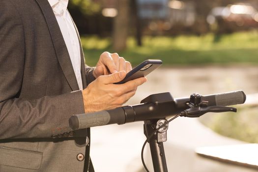 Unknown businessman in classic suit is coming up to an electric scooter and using mobile phone app. Man rents electric scooter. Eco-friendly transportation. Outdoors of business center.