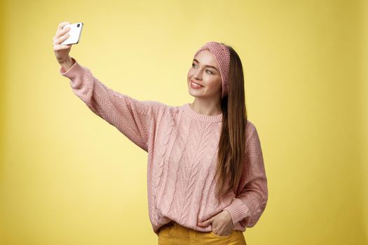 Popular glamour young female internet lifestyle blogger taking selfie on new smartphone extending arm taking picture herself against yellow background smiling at cellphone screen, posing cheeky.