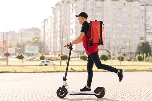 Man courier food delivery with red thermal backpack rides the street on an electric scooter deliver online order client customer. Deliveryman worker employee in red uniform. Excellent delivery.