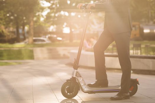 Unknown businessman in classic suit ride on electric mobile scooter. Close up of legs man in suit riding electric scooter in city. Modern eco friendly transport. Fast speed driving electric transport.