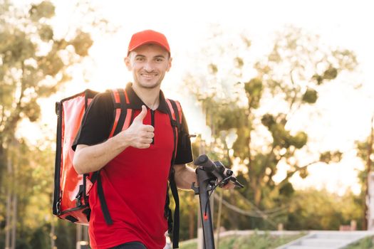 Portrait smiling man courier food delivery standing with thermal backpack, electric scooter looking at camera and showing thumb up. Positive deliveryman worker deliver online order client