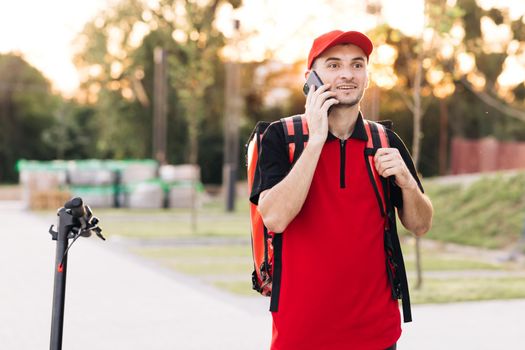 Male courier with isothermal food case box arrives on a electric scooter to the entrance to the house and calls for client. Food delivery guy with red backpack searching delivery addresses.