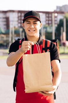 Portrait of delivery man with red uniform holding food bags waiting for customer. Close-up happy young courier is proud of his job smiling standing in the street. Home delivery.