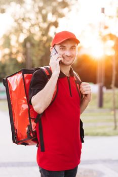 Male courier with isothermal food case box on electric scooter arrives to the entrance to the house and calls for client. Food delivery guy with red backpack deliver orders.