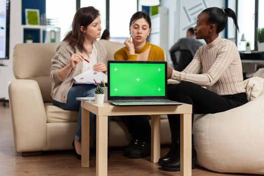 Laptop with chroma key display on table in startup business workplace. African manager discussing talking project statistics with employees on modren office sitting on couch.