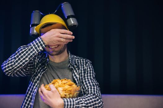 Caucasian man rests at home and watches TV shows or sports news on TV screen. Man with beer helmet on the head eats chips while sitting at home on couch, in evening he watches movies at home on TV.