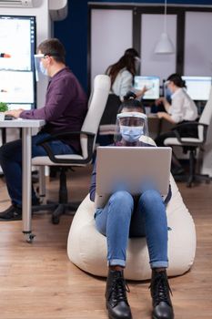 African employee with protection mask against covid-19 pandemic sitting on armchair in middle of office room holding digital tablet. Multiethnic business team working respecting social distance during global pandemic with coronavirus.