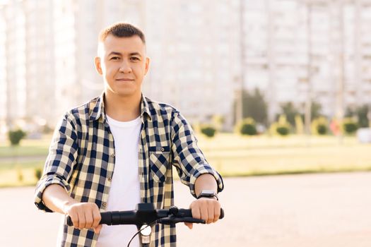 Young man standing in front of city, holding e-scooter. Eco transportation. Modern urban alternative transport. Eco-friendly transportation.