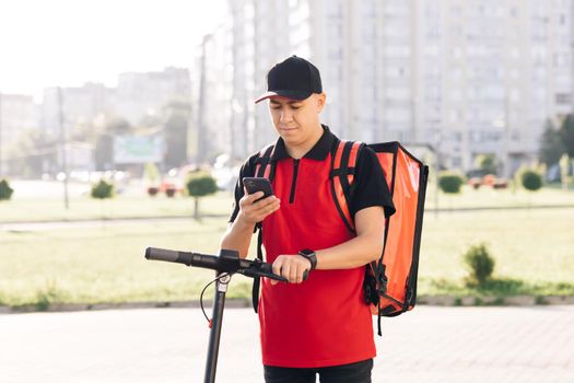 Male courier food delivery with red thermal backpack looks around street with electric scooter uses smartphone navigate. Deliveryman worker employee deliver online order client customer.